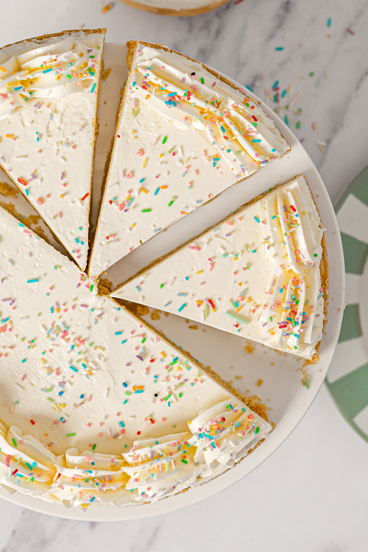 Overhead view of sliced sugar cookie cake with vanilla frosting and rainbow sprinkles on a white plate.