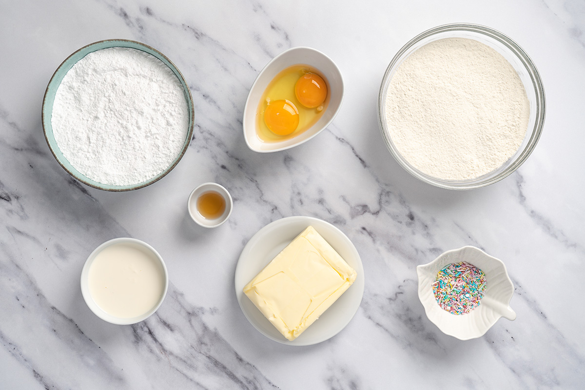 Ingredients for sugar cookie cake laid out on marble: sugar cookie mix, butter, eggs, powdered sugar, vanilla, cream, sprinkles.