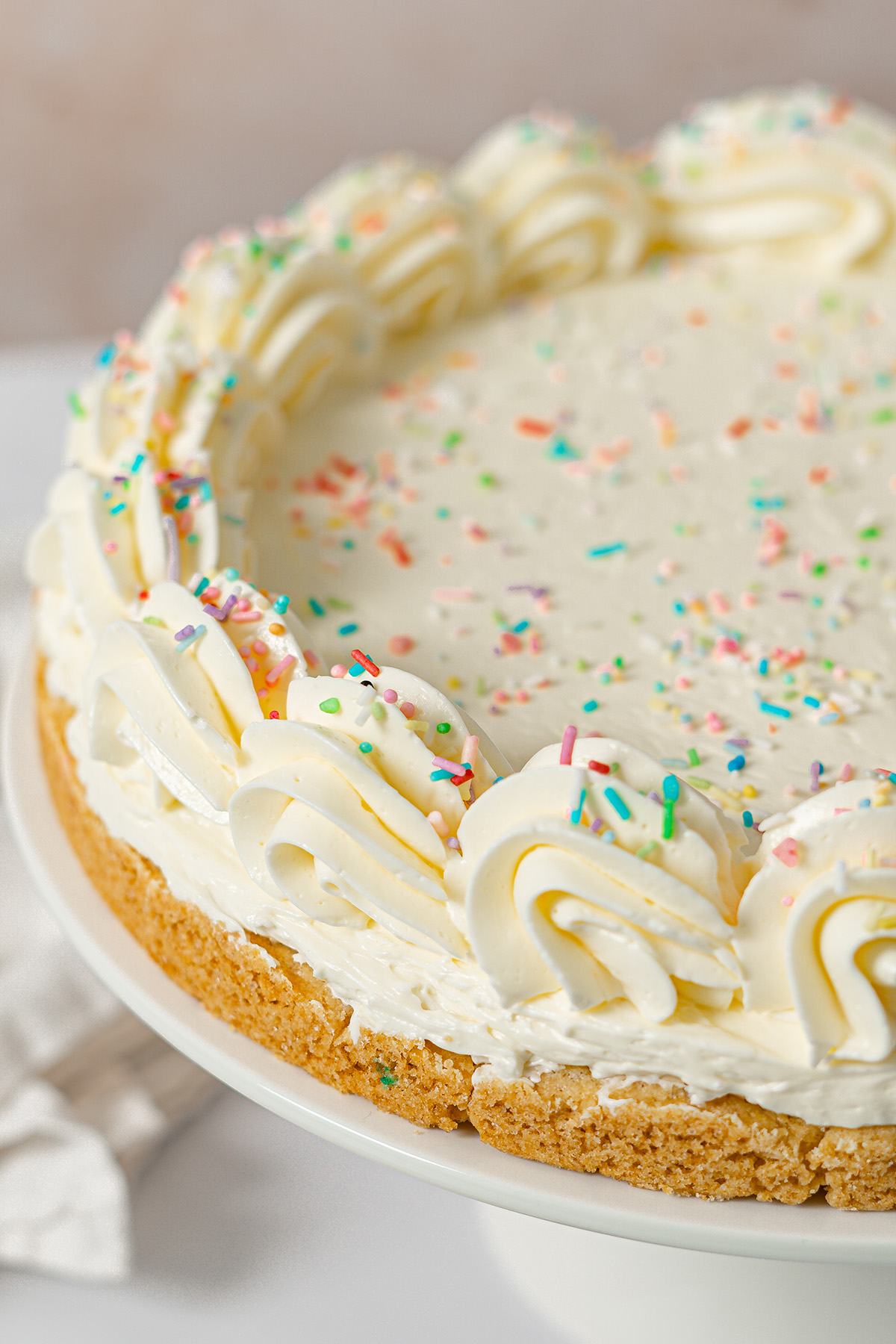 Sugar cookie cake on a white cake stand topped with vanilla frosting swirls and rainbow sprinkles around the edge.