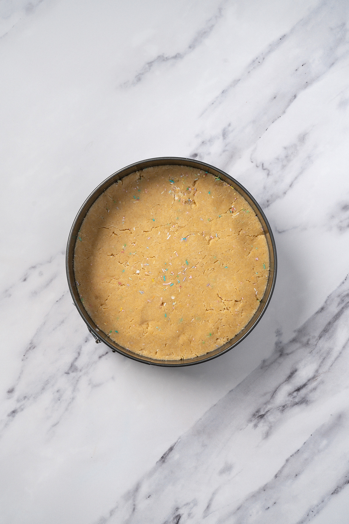 Sugar cookie cake dough pressed evenly into a greased 9-inch springform pan with sprinkles throughout.