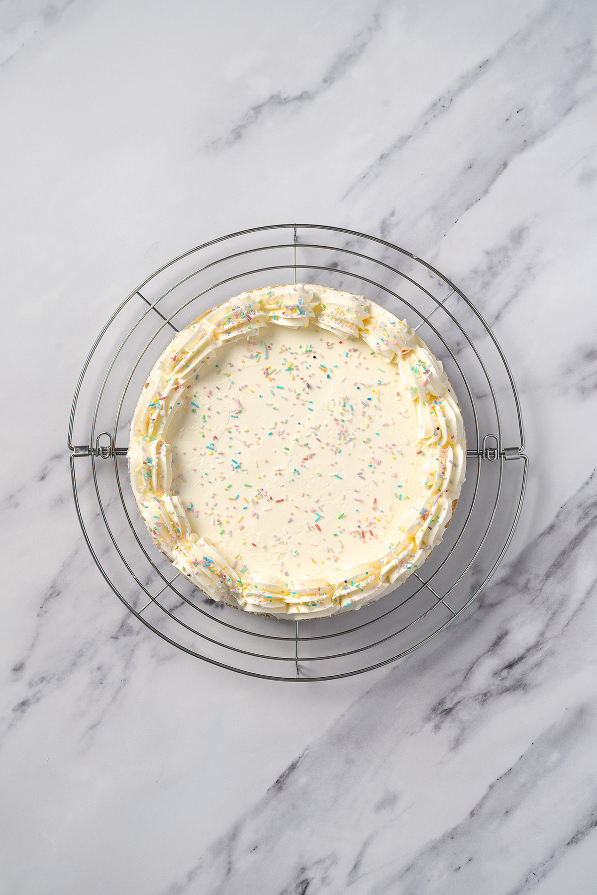 Frosted sugar cookie cake on a wire cooling rack with vanilla buttercream and rainbow sprinkles on top.