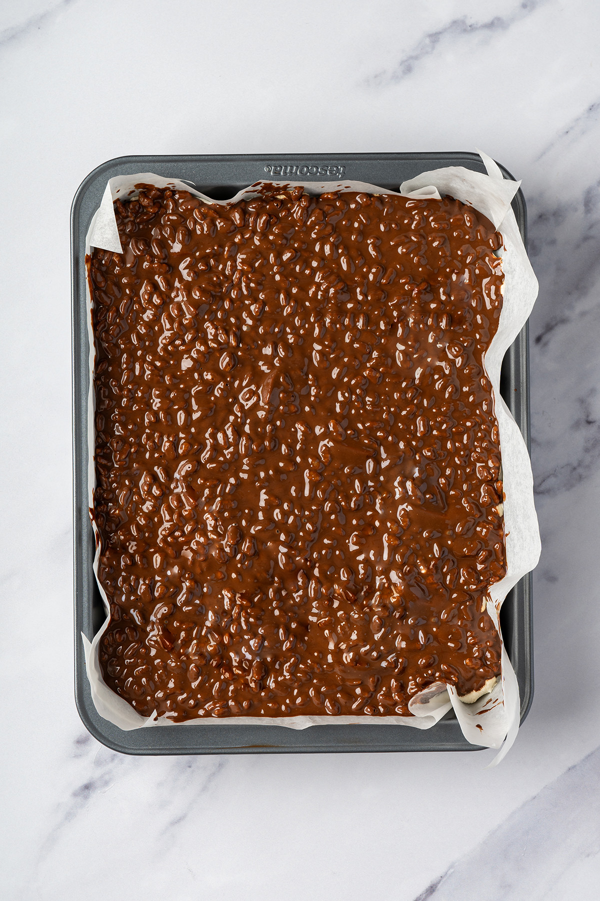 Chocolate peanut butter Rice Krispie topping spread over marshmallow brownies in a parchment-lined pan.