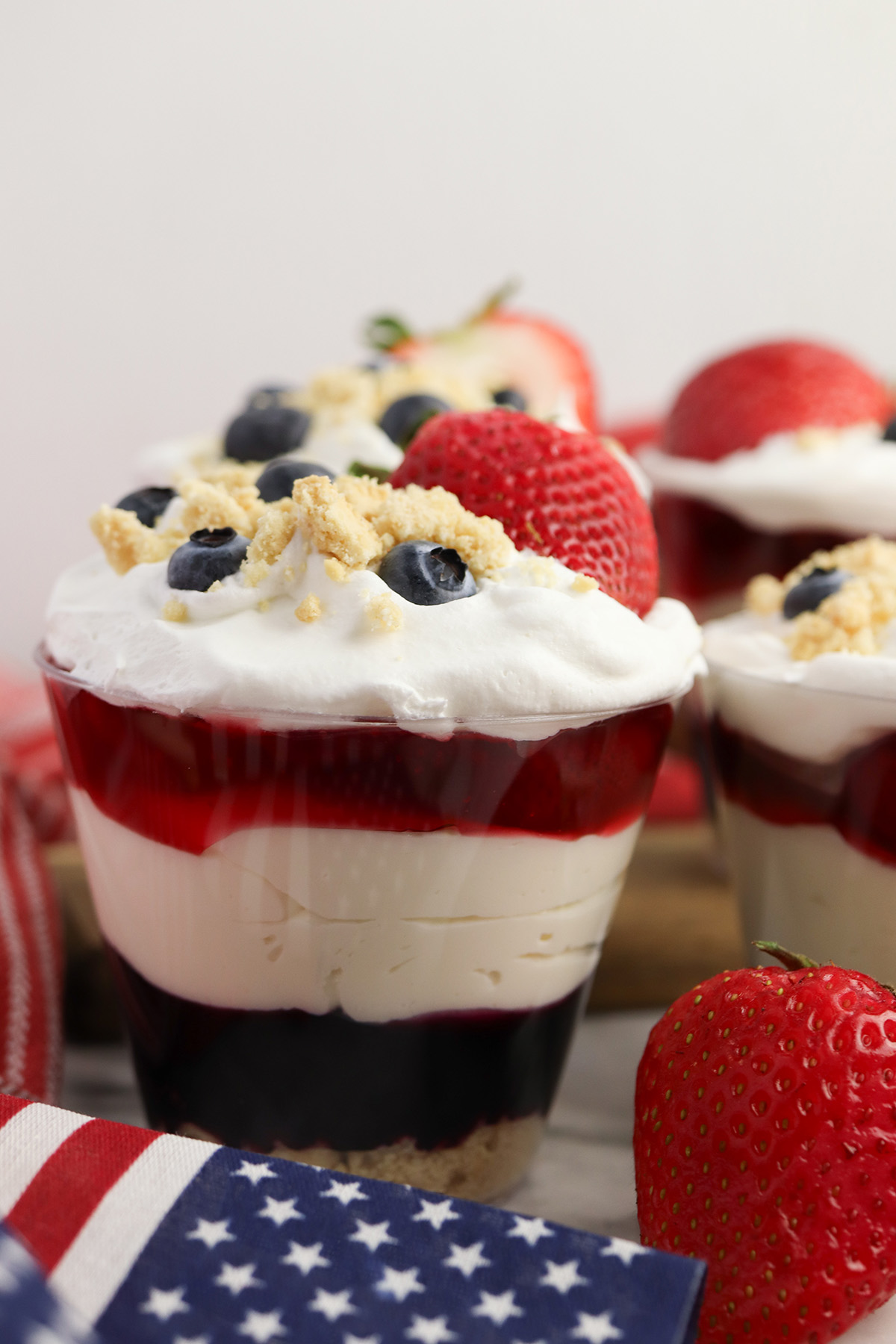 Close up of a patriotic no bake cheesecake cup showing layers of golden Oreo crumbs, blueberry pie filling, creamy cheesecake filling, strawberry pie filling, whipped topping and garnish.
