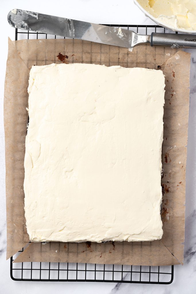 Cooled brownie slab spread with vanilla frosting on parchment paper.