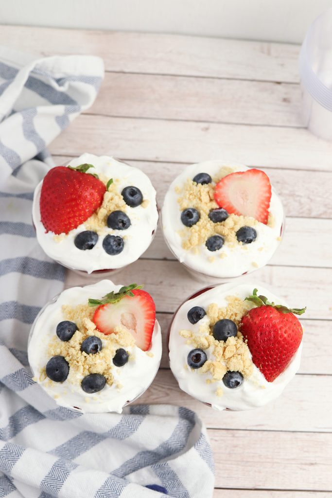 Four patriotic no bake cheesecake cups topped with whipped topping, golden Oreo crumbs, strawberries, and blueberries.