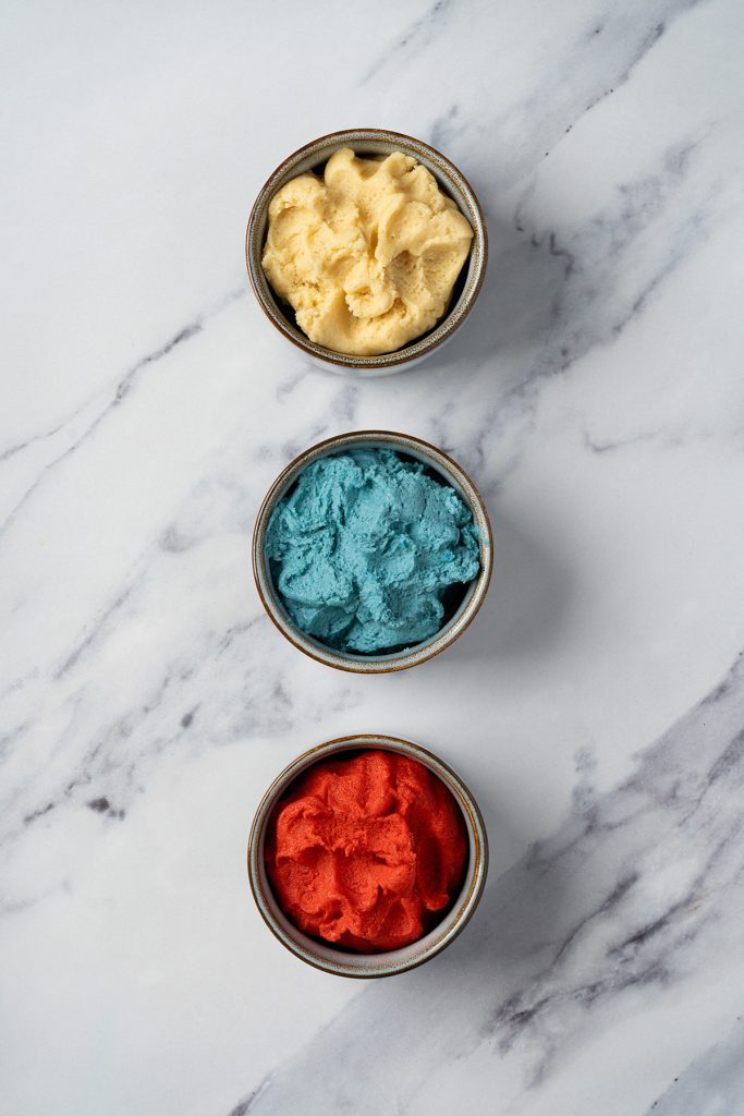 Three bowls of red, blue and plain sugar cookie dough on a marble surface.