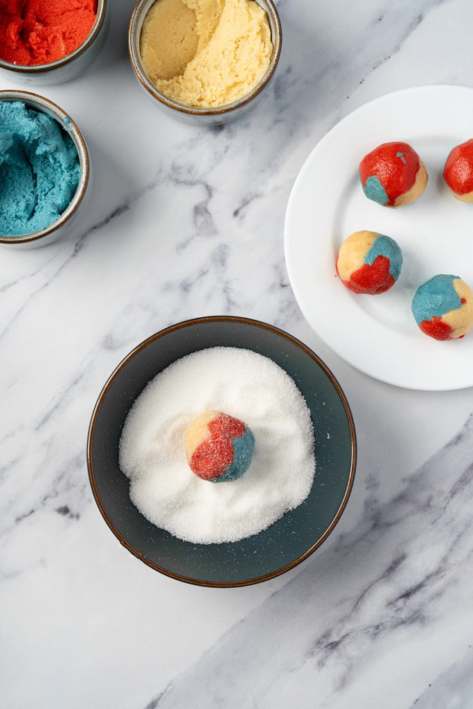 Red, white and blue sugar cookie dough ball being rolled in granulated sugar.