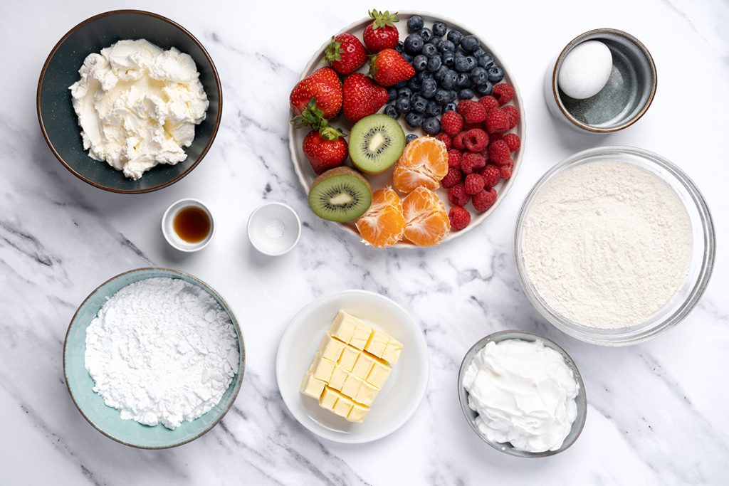 Ingredients for fruit pizza sugar cookie cups with sugar cookie mix, cream cheese, frosting, fresh berries, kiwi, oranges, butter, egg, and powdered sugar.