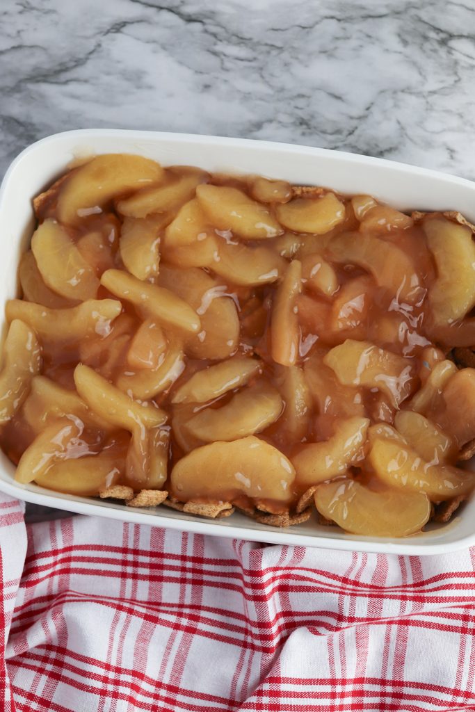 Apple pie filling spread over Cinnamon Toast Crunch cereal in a white baking dish for apple dump cake.
